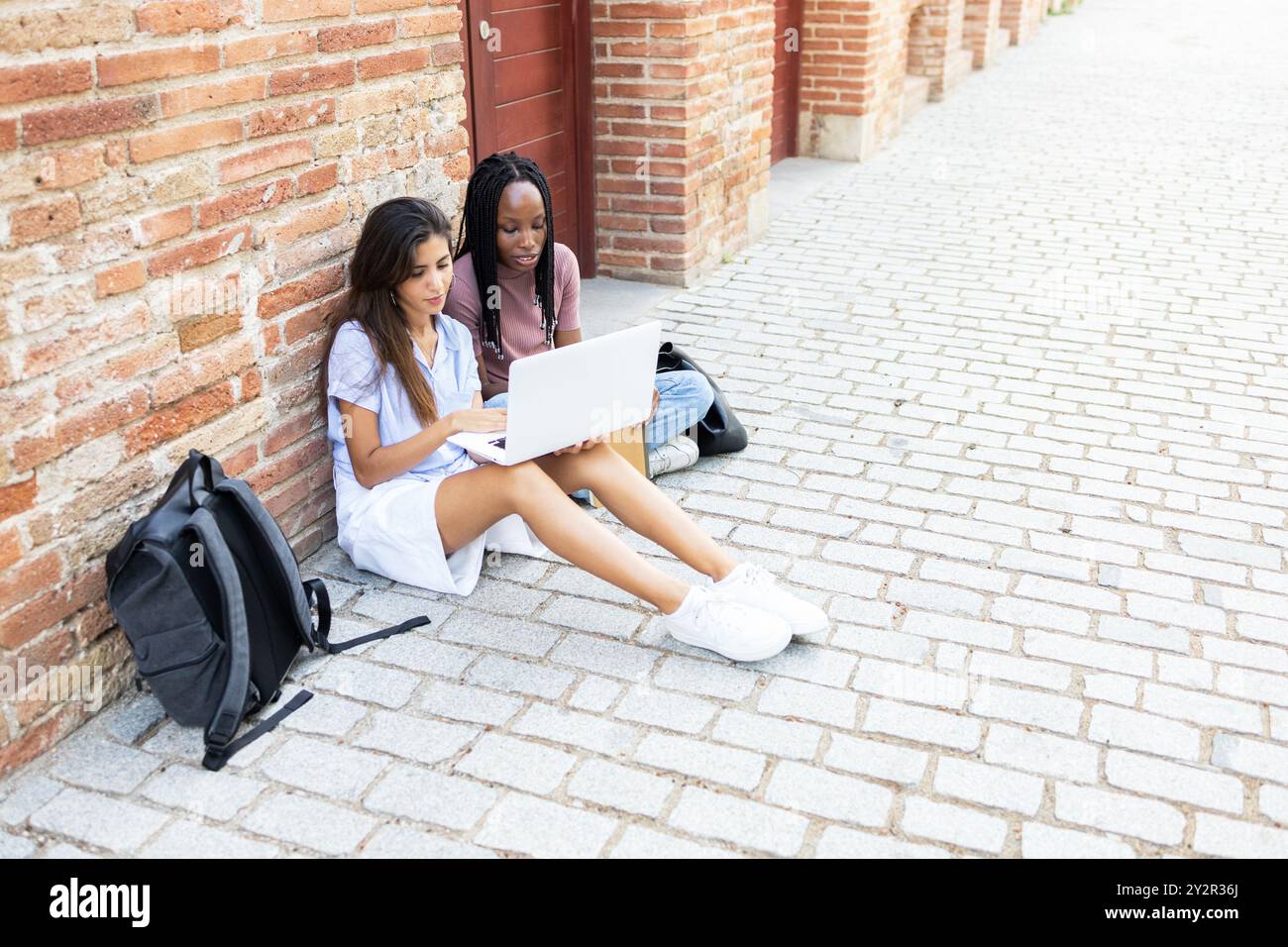 Due giovani donne, sedute su un marciapiede di mattoni e che studiano da un laptop, con gli zaini accanto, simboleggiano l'apprendimento collaborativo Foto Stock