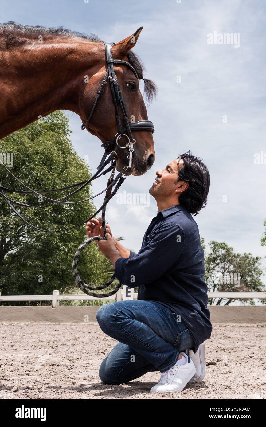 Uno sposo attento si inginocchia delicatamente per tenere e interagire con un cavallo bruno in un centro equestre, dimostrando un legame di fiducia e affetto sotto una clea Foto Stock