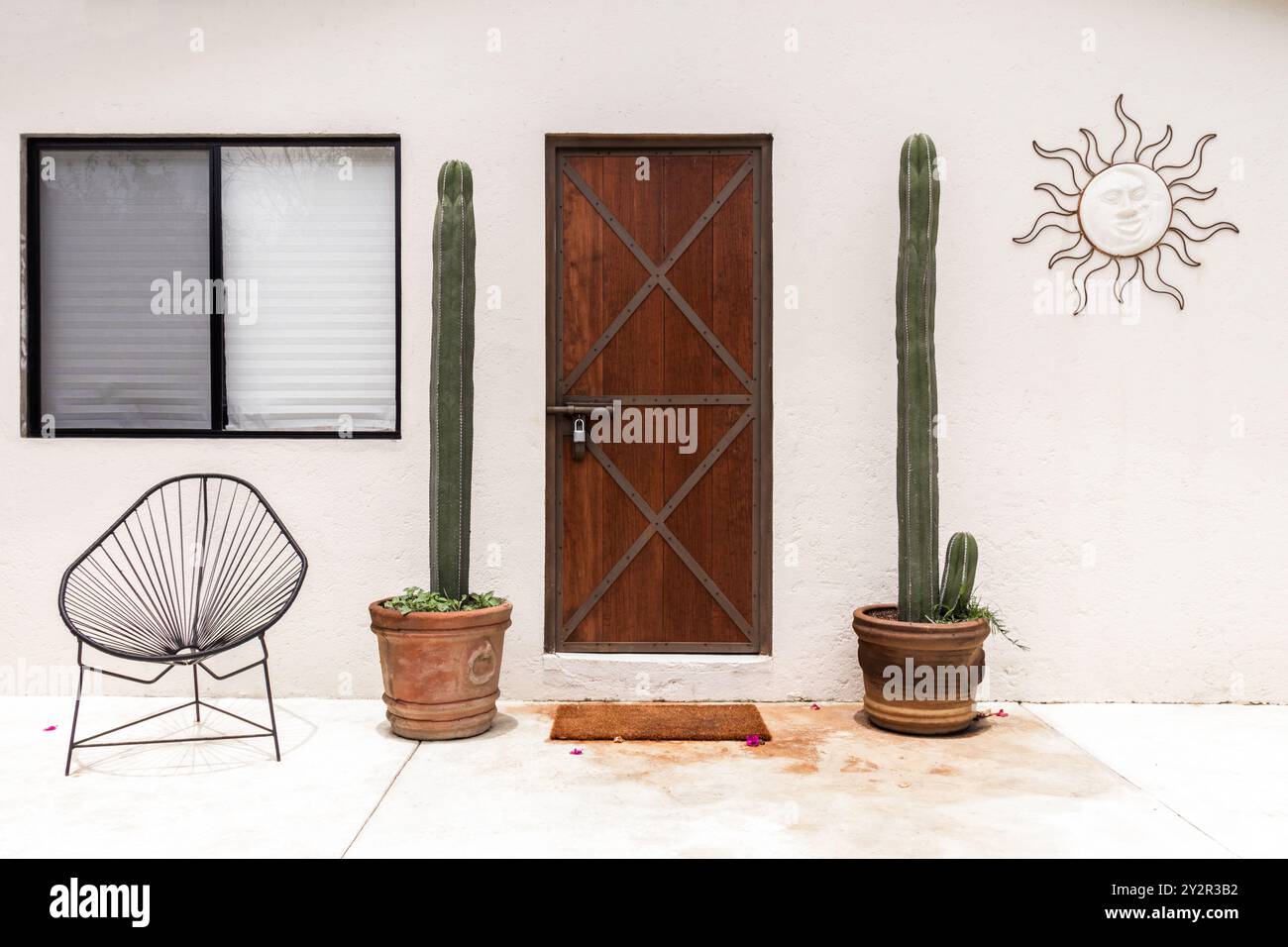 Questa immagine mostra un patio esterno in stile sud-occidentale caratterizzato da una sedia geometrica in filo metallico e una porta in legno rustico, incorniciata da alti cactus in terracotta po Foto Stock