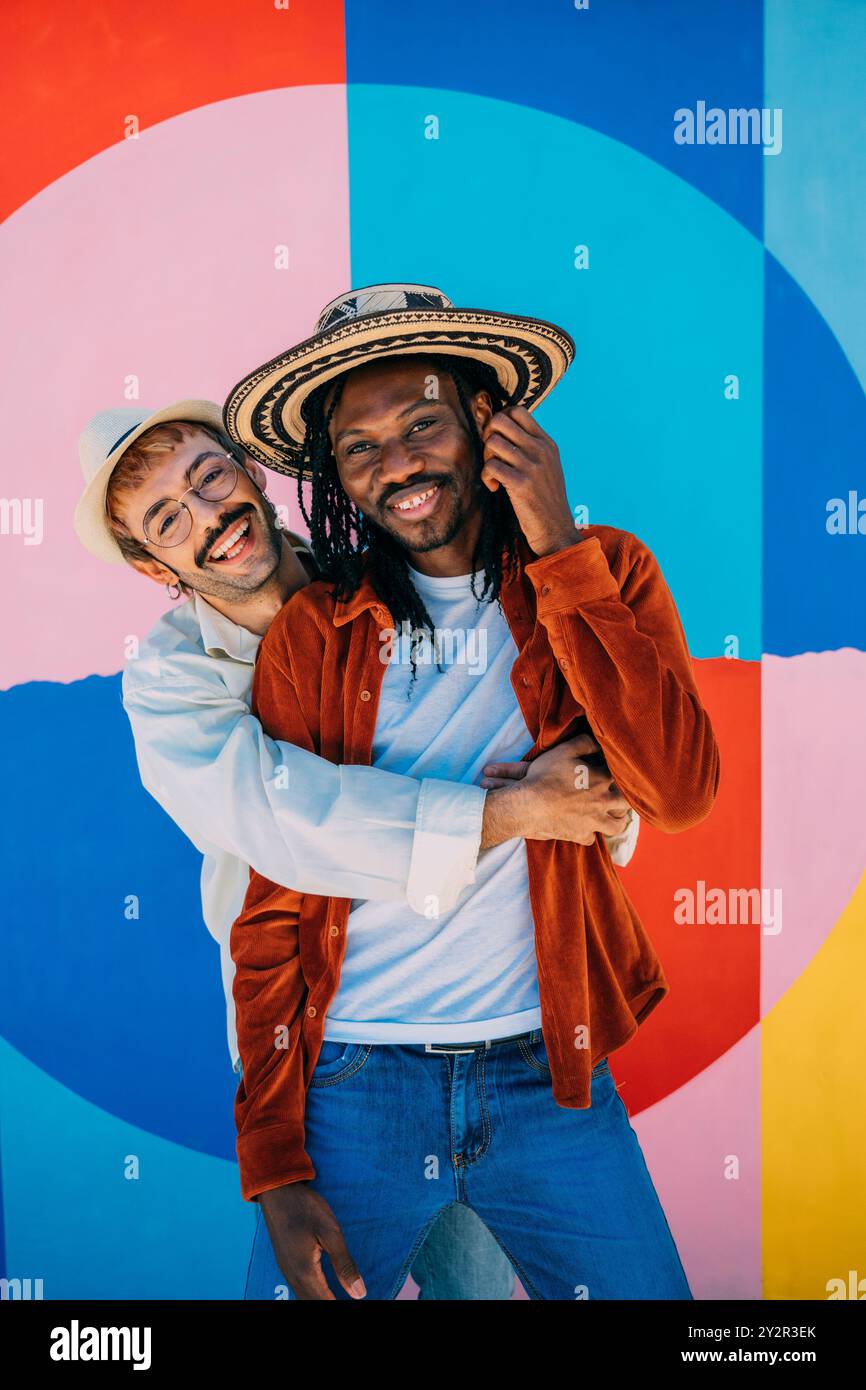 Una coppia gay, tra cui un uomo di colore, posa felicemente davanti a uno sfondo vibrante e colorato che irradia gioia e amore per gli uomini, uno che indossa un sombrero e il Foto Stock