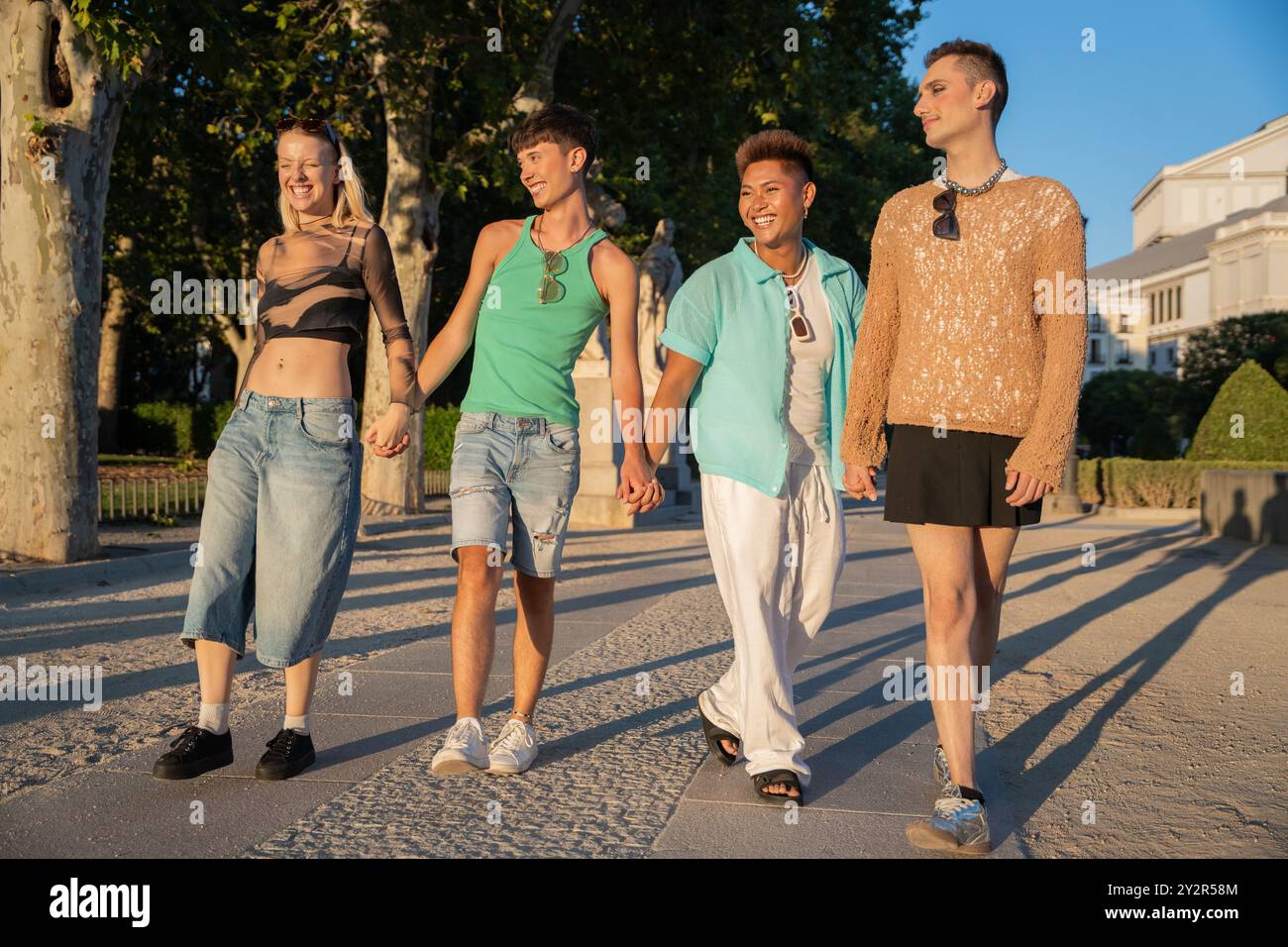Un gruppo eterogeneo di amici LGBTQIA+ che si tengono per mano e sorridono durante una piacevole passeggiata in un parco illuminato dal sole, simboleggiando l'amicizia e l'inclusione. Foto Stock