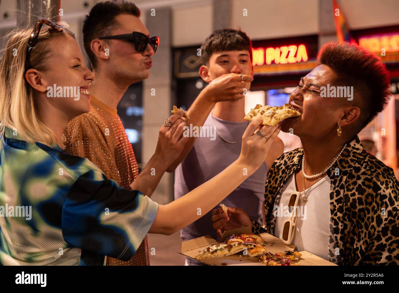 Gli allegri amici LGBTQIA+ si godranno un momento di pizza informale insieme, mostrando diversità e comunità in un vivace ambiente urbano. Foto Stock