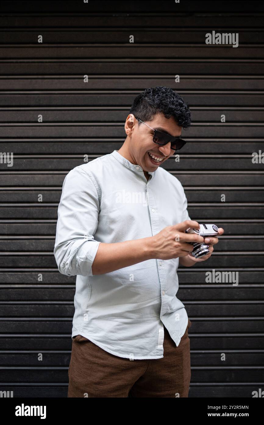 Un allegro giovane latino in un elegante abbigliamento casual ride mentre maneggia una macchina fotografica d'epoca, davanti a uno sfondo di metallo scuro. Foto Stock