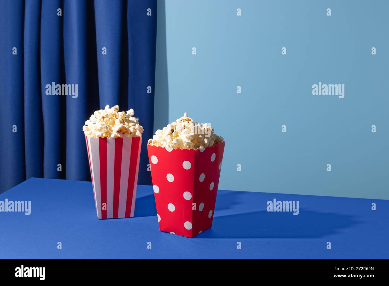 Due contenitori di popcorn, uno rosso con strisce bianche e l'altro rosso con pois bianchi, siedono su un tavolo blu contro una tenda blu coordinata Foto Stock