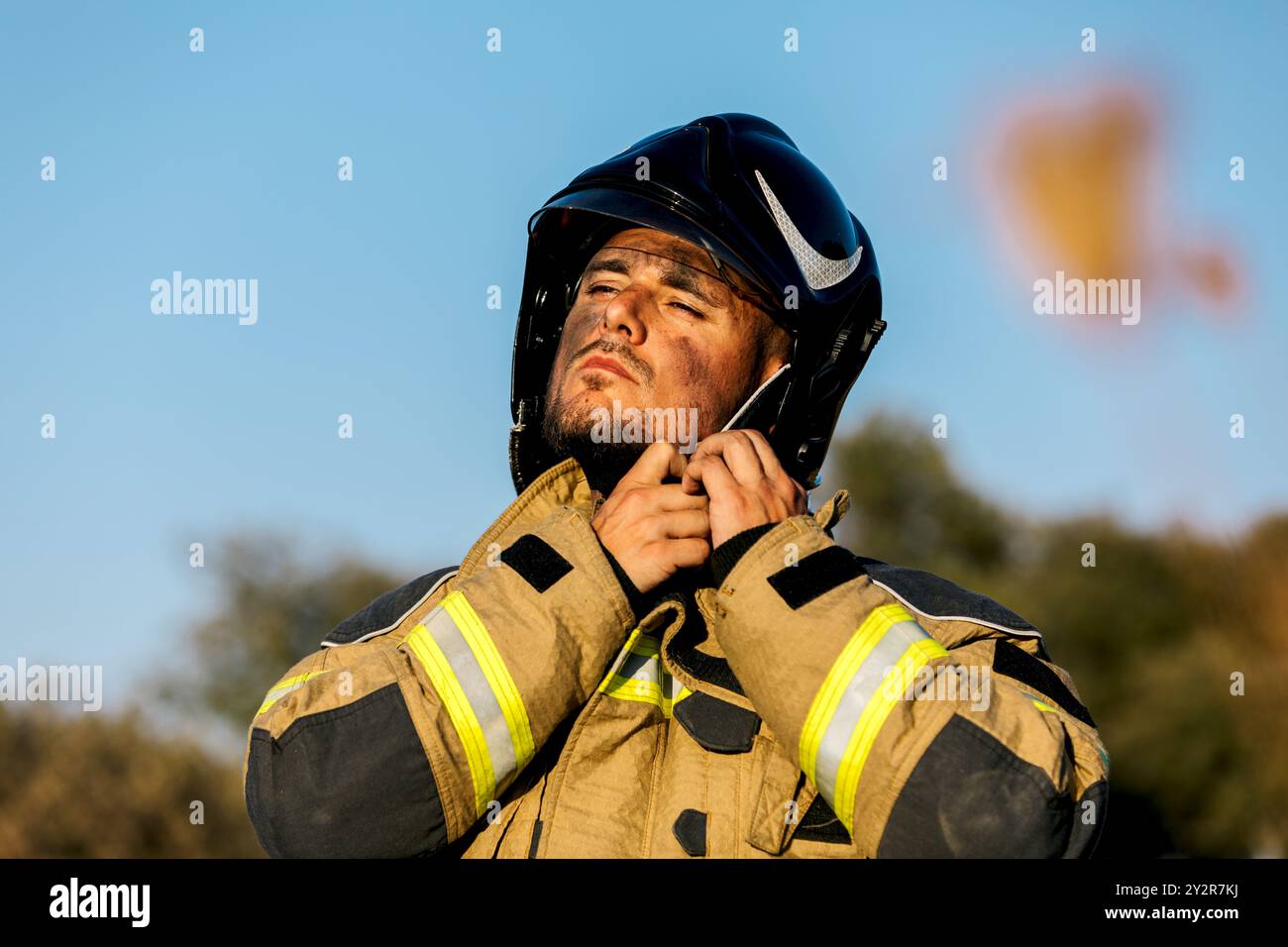 Un vigile del fuoco con equipaggiamento protettivo regola il suo casco mentre si concentra su un compito, con un fuoco boschivo sfocato visibile sullo sfondo che l'immagine cattura Foto Stock