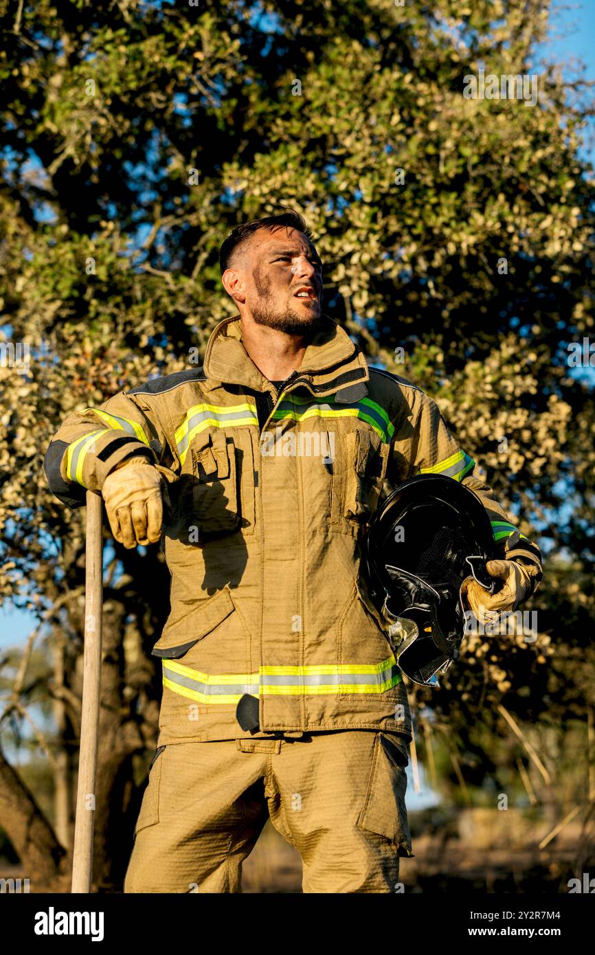 Un robusto vigile del fuoco, vestito con un equipaggiamento protettivo completo con strisce riflettenti, tiene il casco mentre si trova in una foresta bagnata dalla luce del sole, con spettacoli Foto Stock