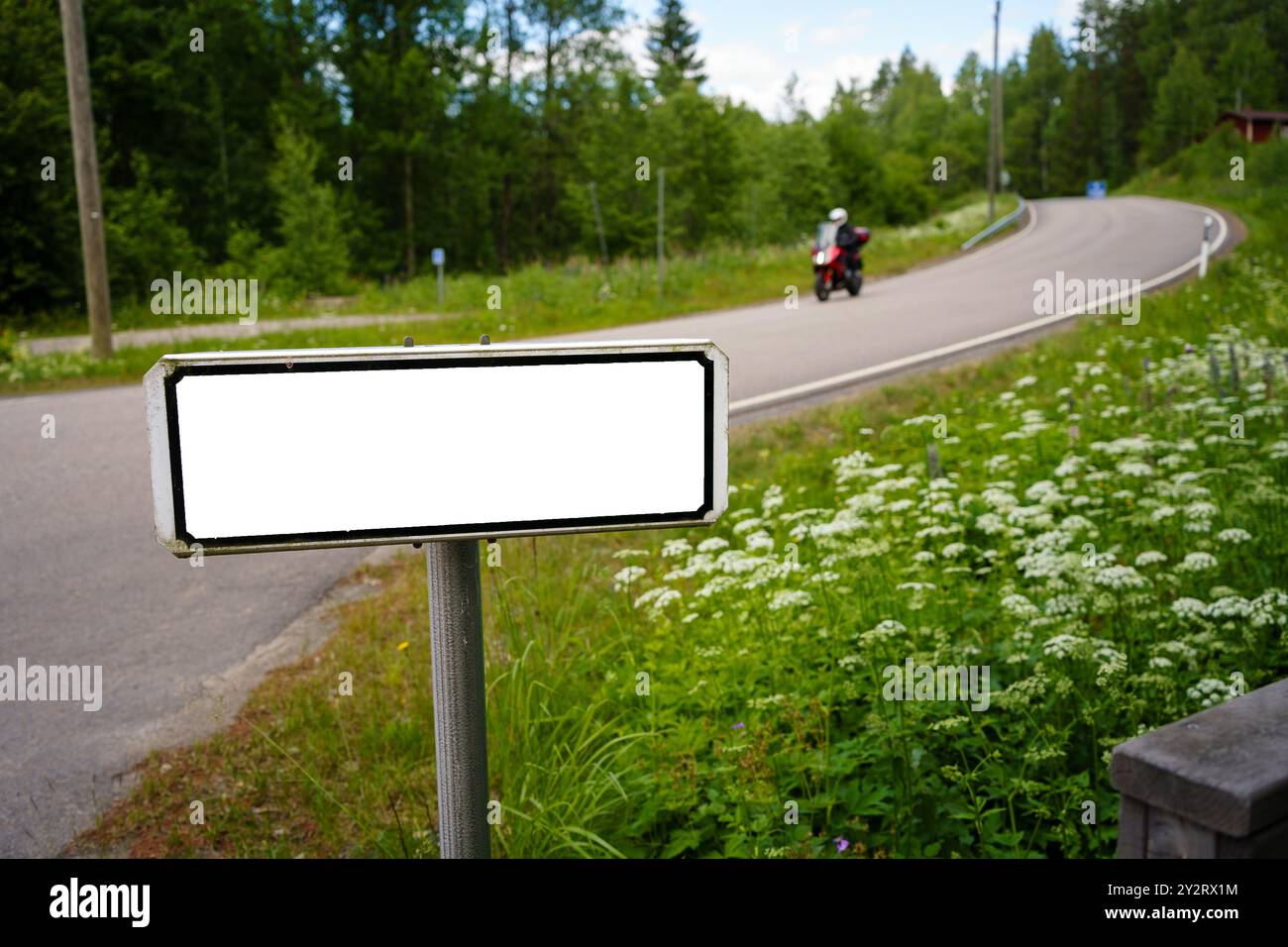 Un cartello stradale vuoto vicino a una tortuosa strada di campagna, con una motocicletta in lontananza Foto Stock