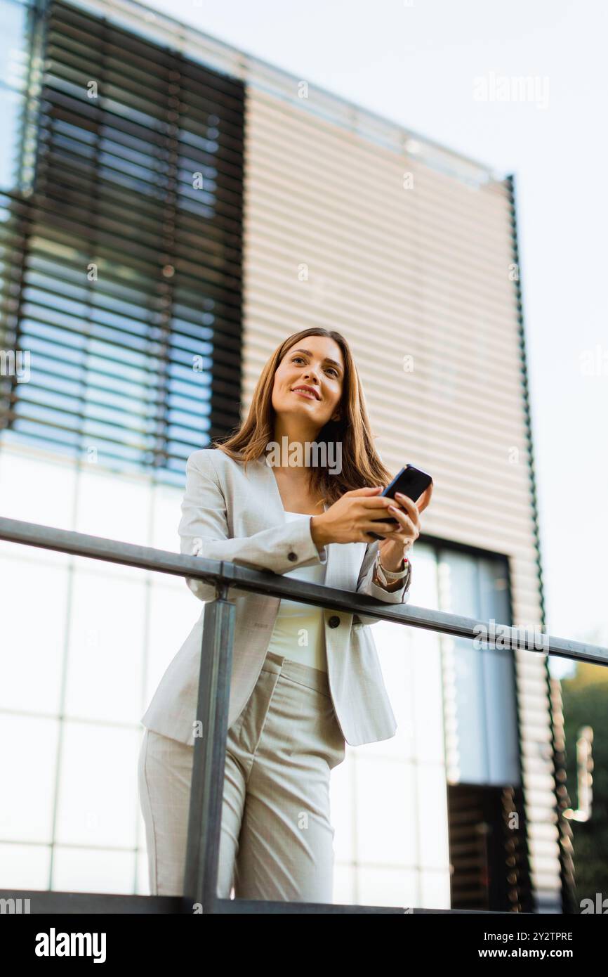 Allegra donna d'affari in piedi su un balcone, impegnata con il suo smartphone su uno sfondo di architettura moderna, immersa nella calda luce del sole e Foto Stock