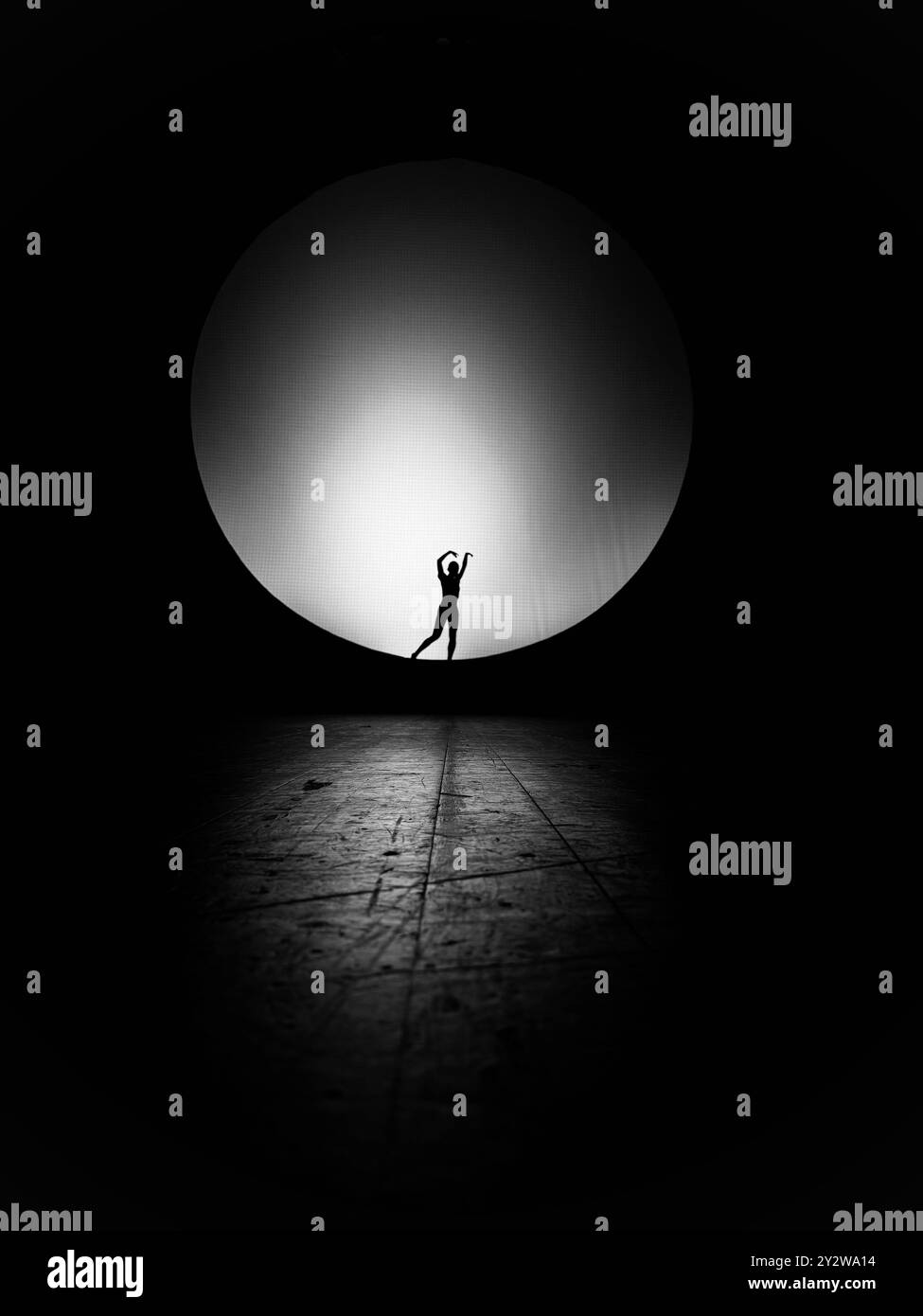 Silhouette di una ballerina che si esibisce sul palco con una grande luce circolare sullo sfondo. Foto Stock