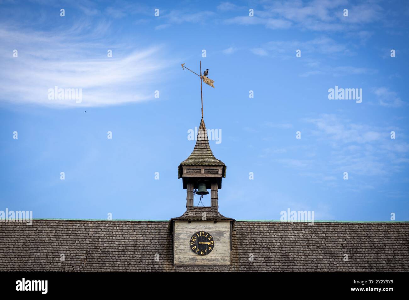 Una vista ravvicinata di una torre dell'orologio con una paletta meteorologica in cima, su un cielo blu con nuvole mostruose. La torre è caratterizzata da una campana e da una spir decorativa Foto Stock