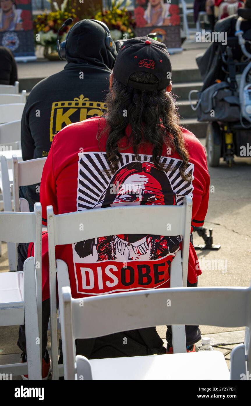 Un uomo siede al programma del giorno indossando una camicia rossa di disobbedienza durante la giornata d'azione delle persone indigene scomparse e uccise al campidoglio. Foto Stock