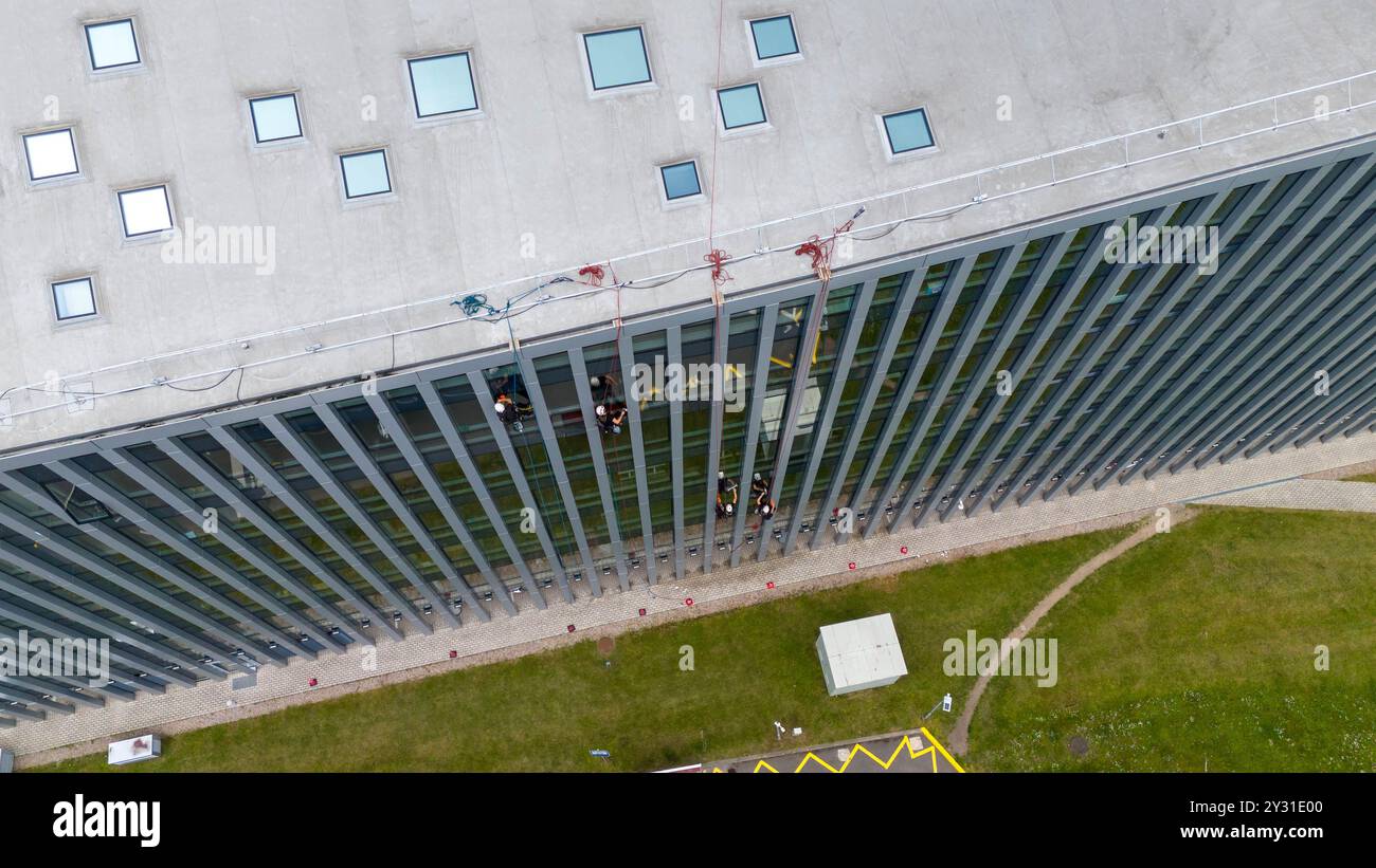 Operai che puliscono le finestre di un moderno edificio in vetro, sospeso su corda. Presenta una struttura alta con superfici riflettenti Foto Stock