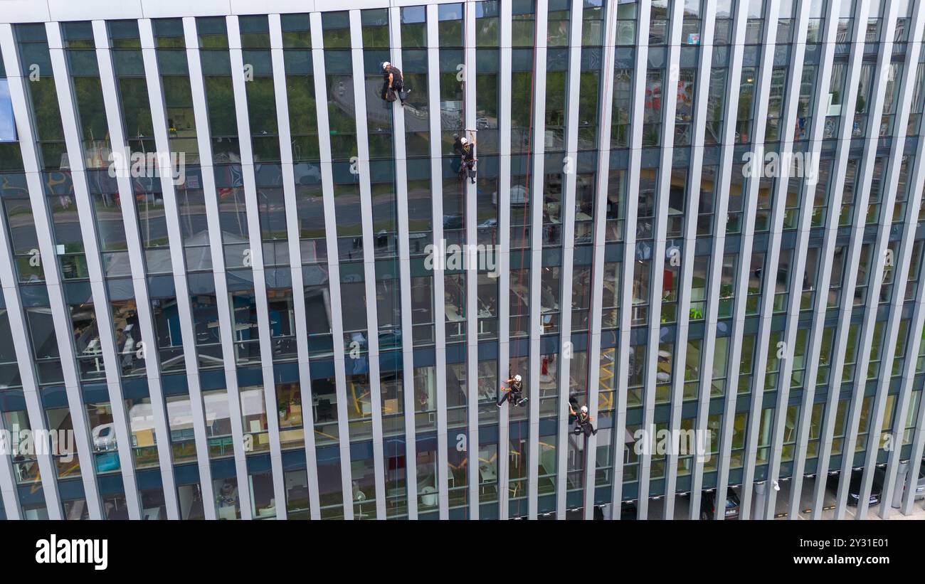 Operai che puliscono le finestre di un moderno edificio in vetro, sospeso su corda. Presenta una struttura alta con superfici riflettenti Foto Stock