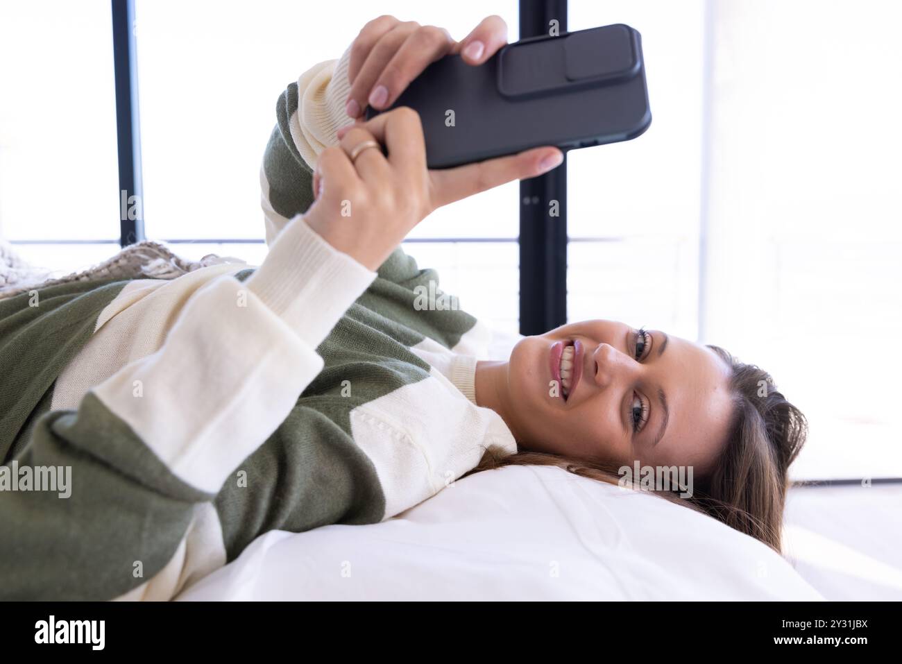 A casa, sdraiata sul letto, giovane donna sorridente e che usa lo smartphone per rilassarsi Foto Stock
