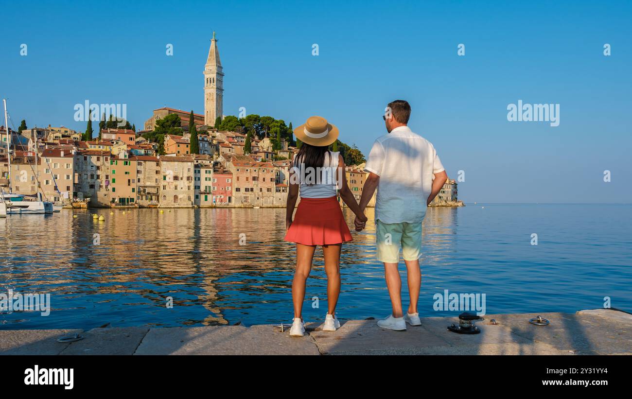 Una coppia si trova mano nella mano sul lungomare, ammirando la pittoresca Rovigno, Croazia. Gli edifici vibranti e la torreggiante torre dell'orologio riflettono il beau Foto Stock