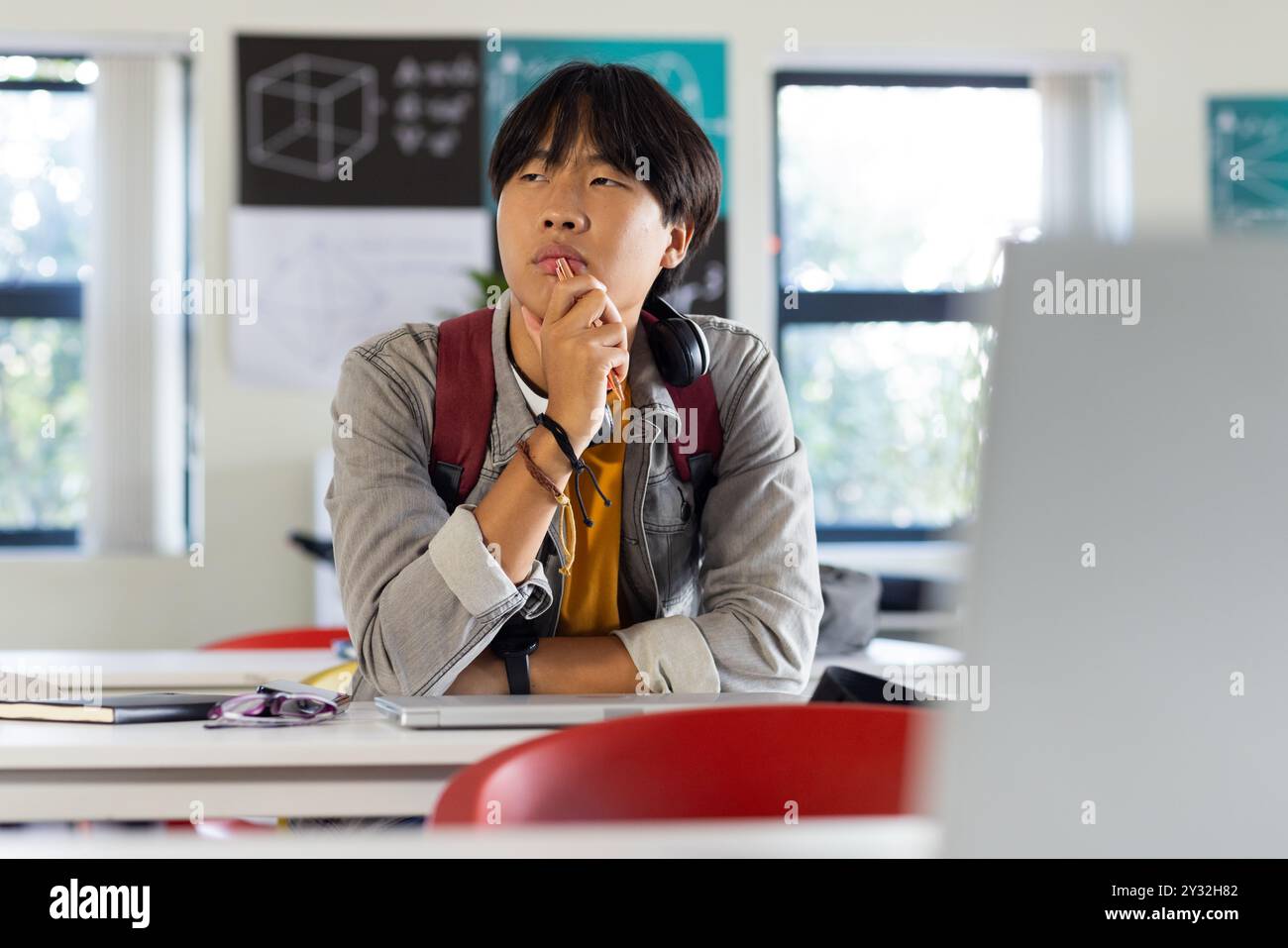 A scuola, adolescente asiatico seduto alla scrivania pensando, con libri e computer portatile nelle vicinanze Foto Stock