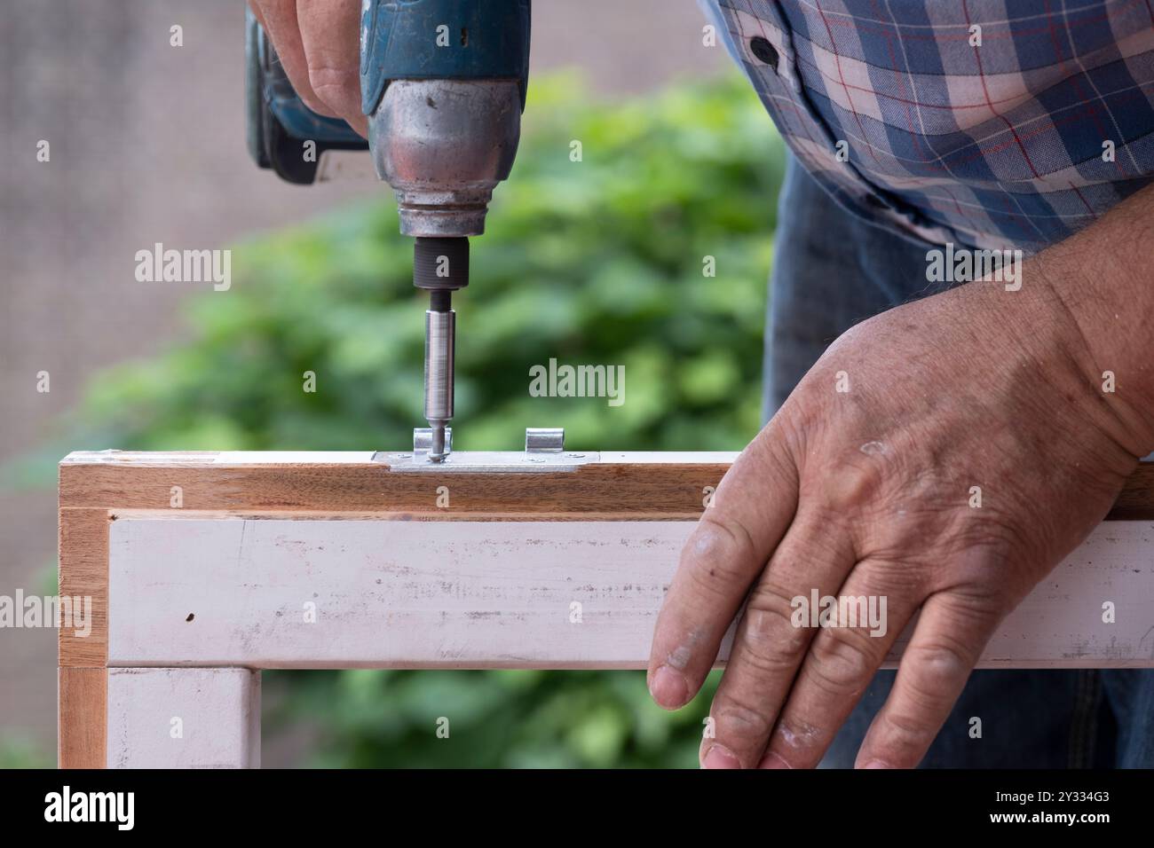 Carpentiere con cacciavite automatico gira una vite in una nuova cerniera di una vecchia finestra in legno. Concetto di ristrutturazione Foto Stock