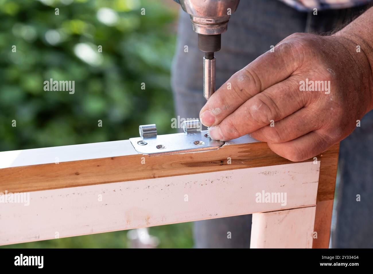 Carpentiere con cacciavite automatico gira una vite in una nuova cerniera di una vecchia finestra in legno. Concetto di ristrutturazione Foto Stock