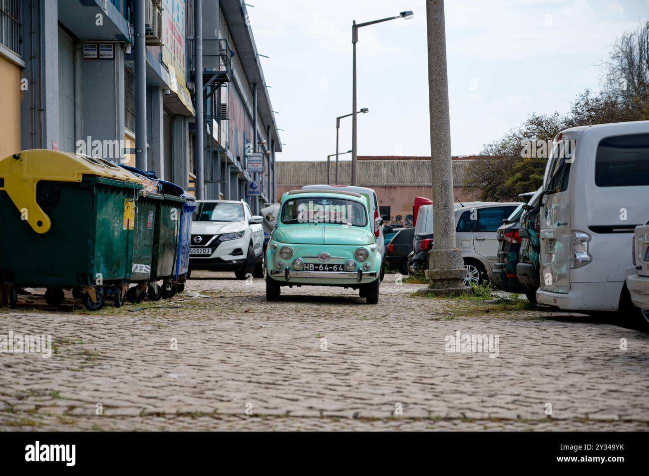 Una Fiat 500 d'epoca verde menta è parcheggiata su una strada acciottolata, circondata da auto e cassonetti moderni Foto Stock