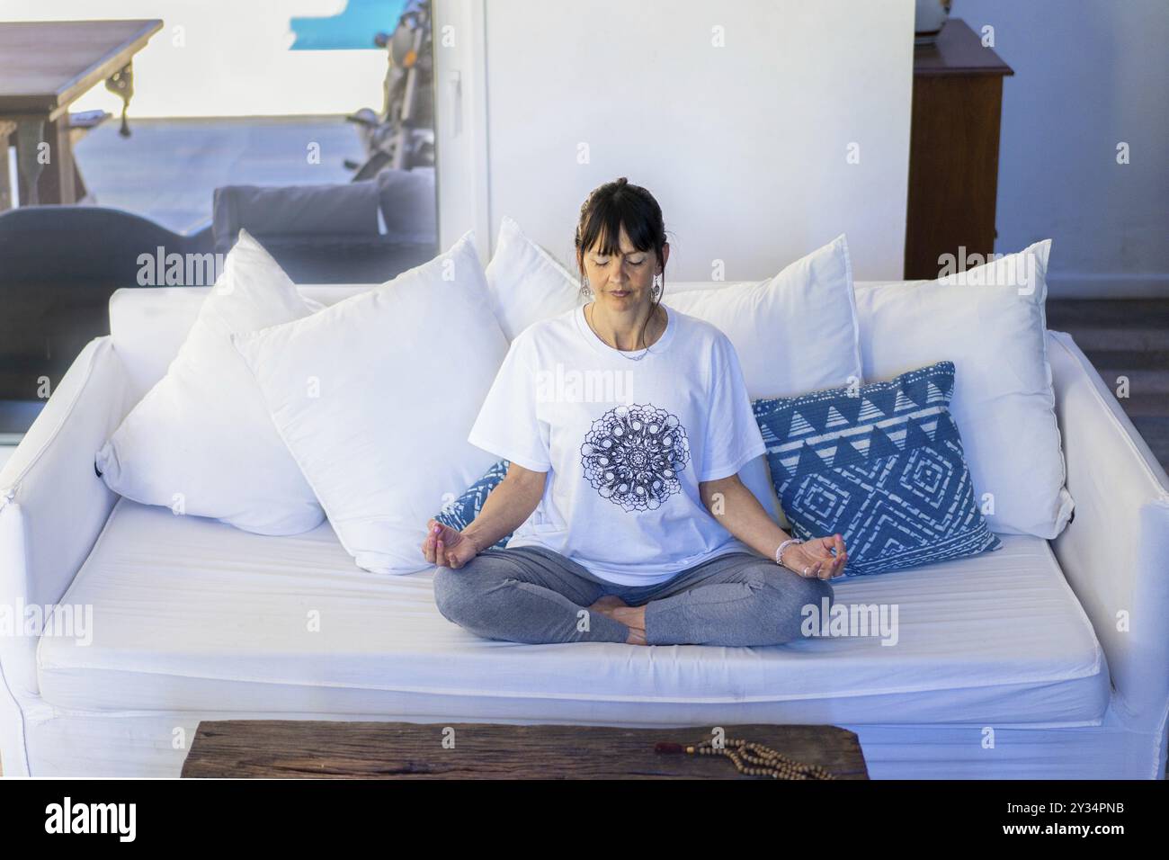 Vista ad angolo alto di una donna di mezza età seduta sul divano a meditare a casa, mani in Gyan Mudra, con bangs e capelli tirati indietro Foto Stock