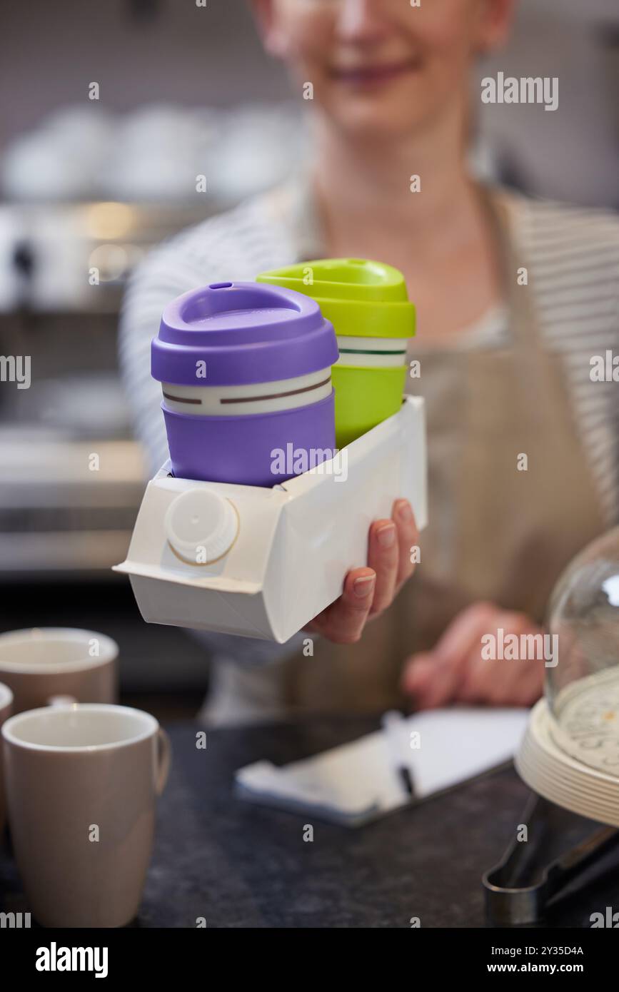 Primo piano di una lavoratrice al caffè che serve caffè o bevande calde in tazze riutilizzabili sostenibili riutilizzando il cartone del latte come portabicchieri Foto Stock
