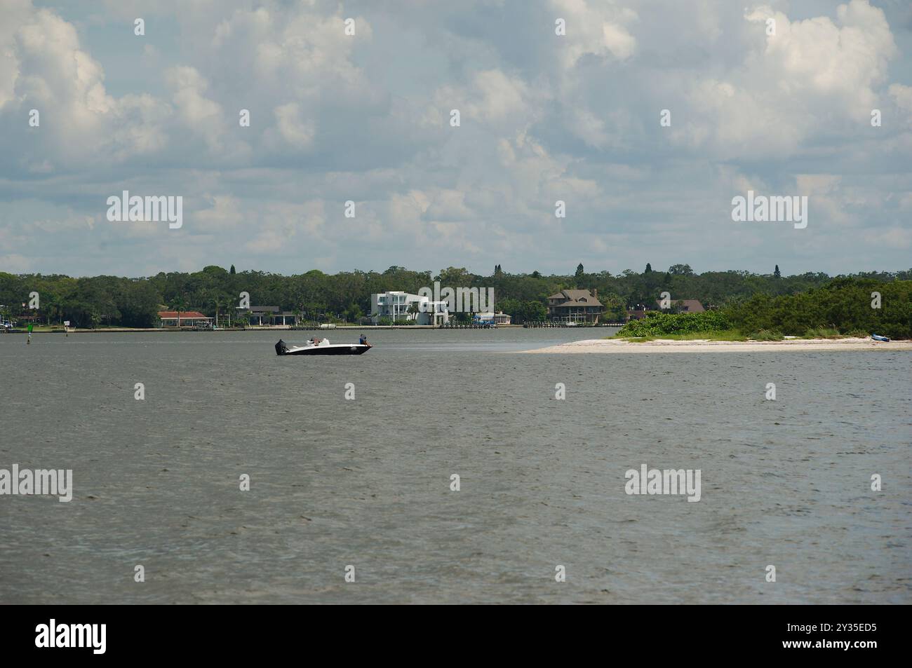 Ampia vista su una spiaggia verso l'acqua della baia con una piccola barca a motore vicino ad una piccola isola di sabbia. Alberi. Cielo azzurro e nuvole bianche luminose e piccolo W Foto Stock