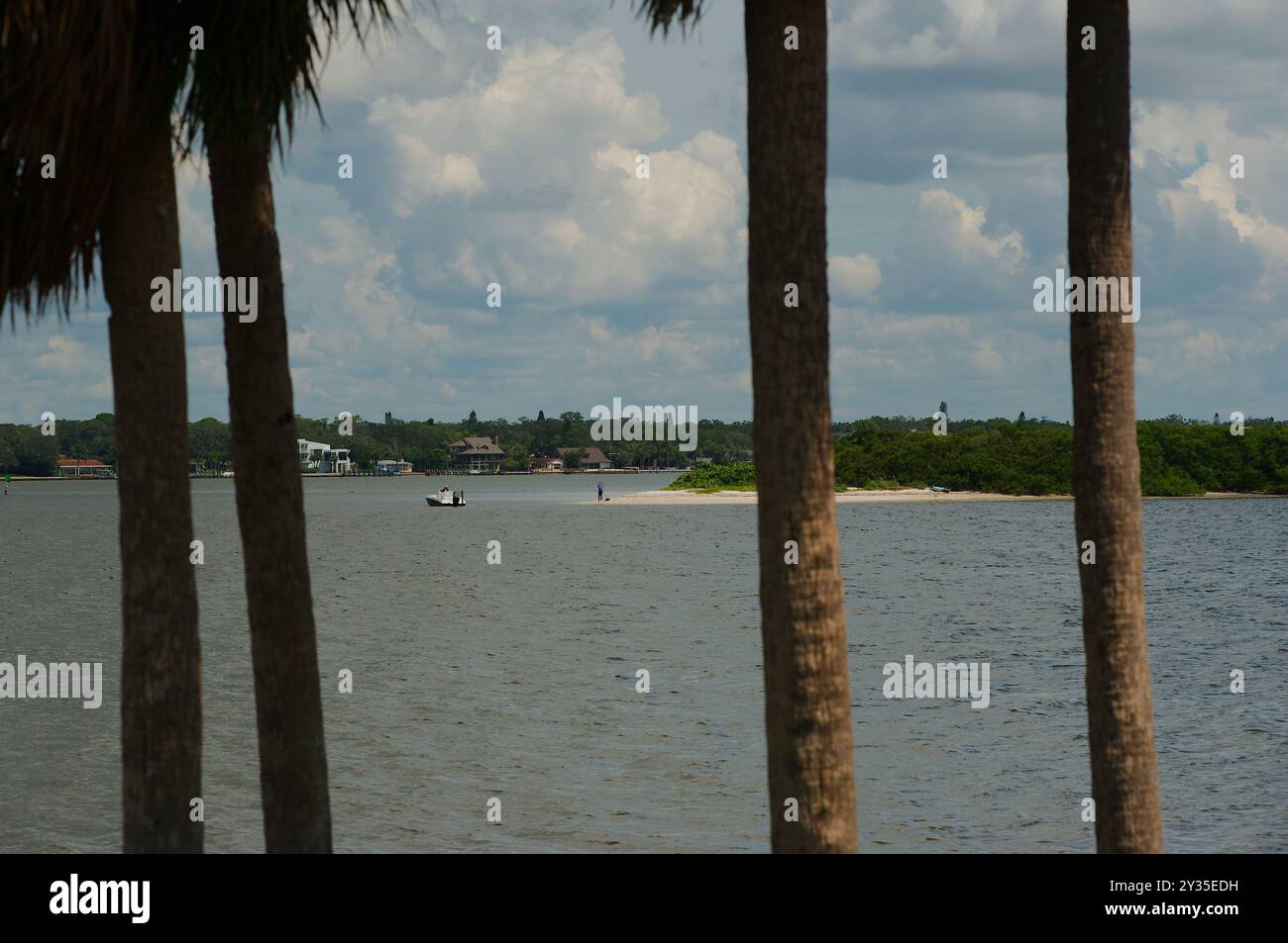 Ampia vista incorniciata tra quattro palme su una spiaggia verso l'acqua della baia con una piccola barca vicino a una piccola isola di sabbia. Alberi. Cielo blu e luminoso Foto Stock