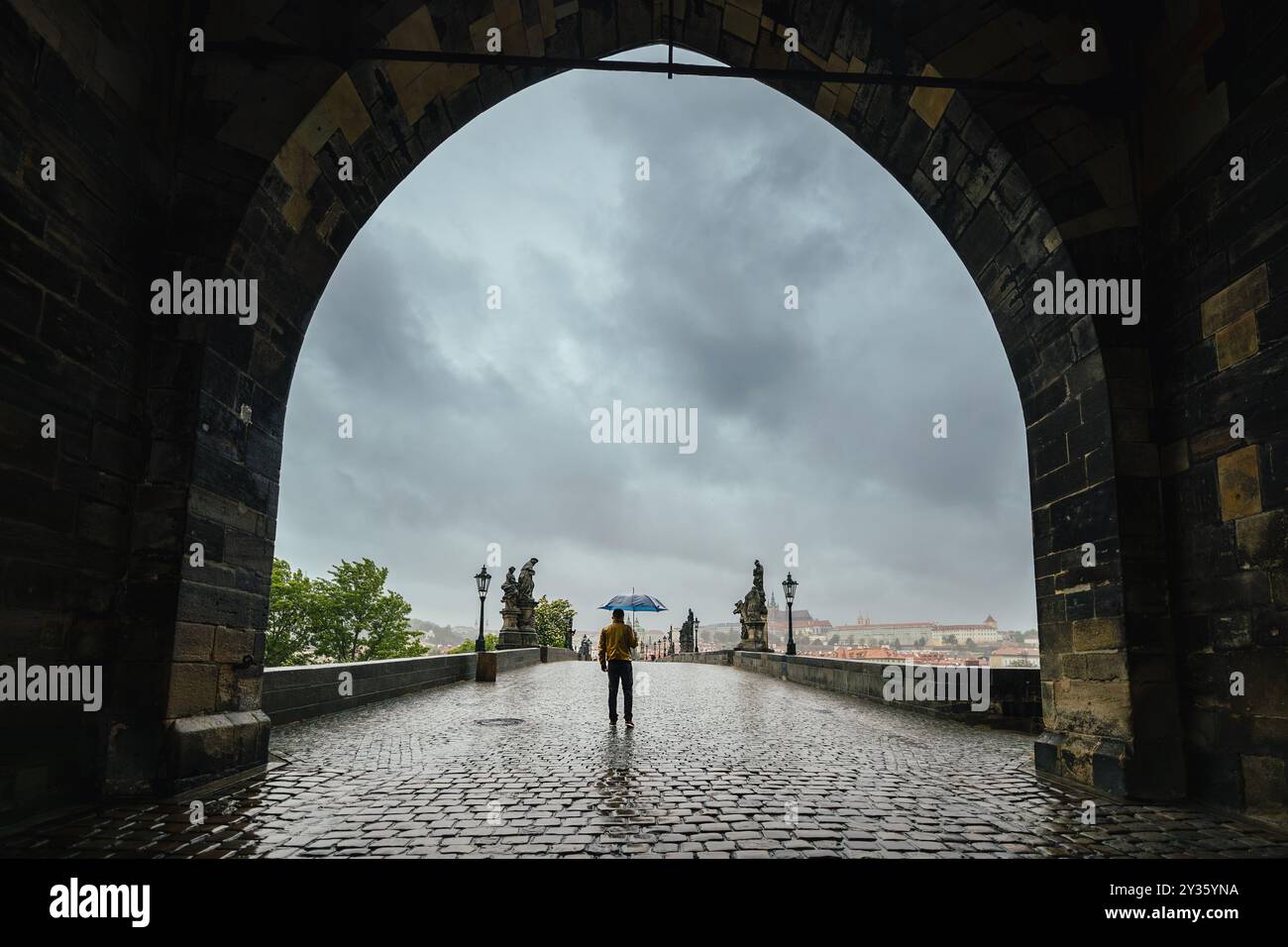 Uomo solitario con ombrello nella città vecchia sotto la pioggia. Vista posteriore della persona che cammina attraverso l'antica porta del Ponte Carlo a Praga in una giornata cupa. Foto Stock