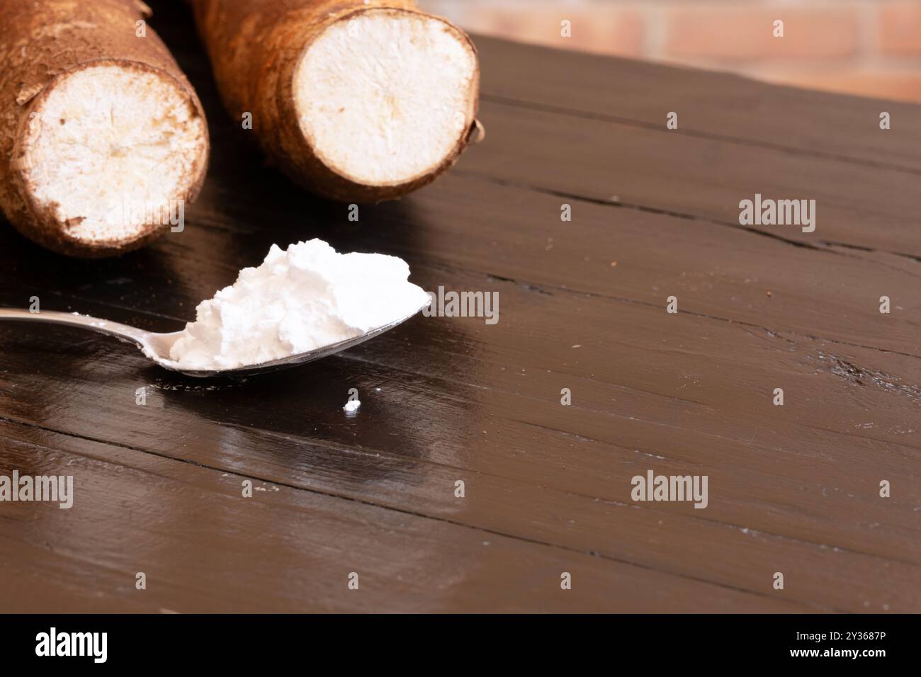 Un cucchiaio con amido di manioca in primo piano e radici di manioca sullo sfondo sopra un tavolo di legno. Foto Stock