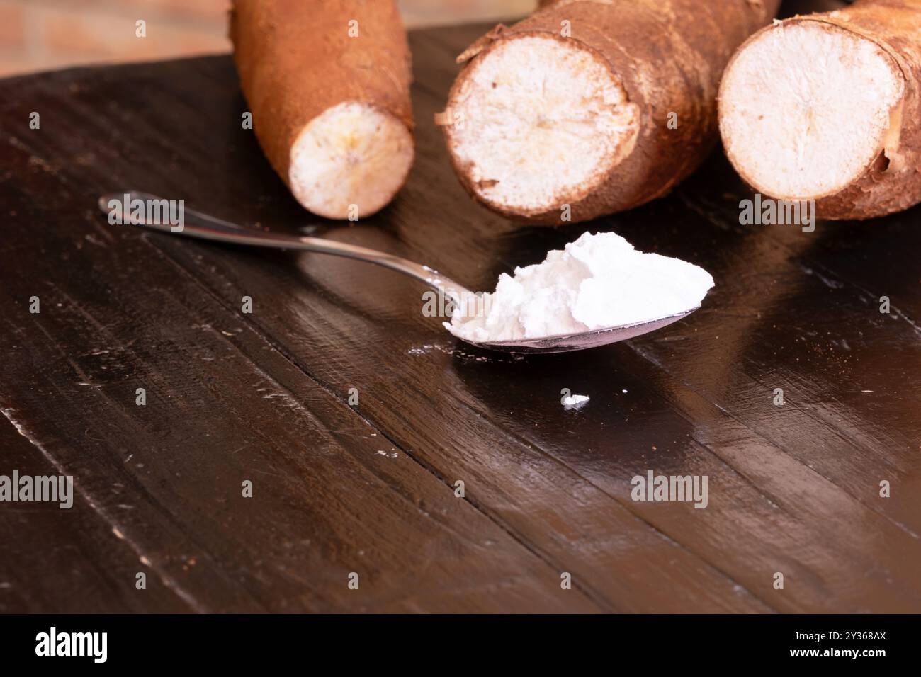 Un cucchiaio pieno di farina di tapioca in primo piano e radici di manioca sullo sfondo sopra un tavolo di legno. Foto Stock
