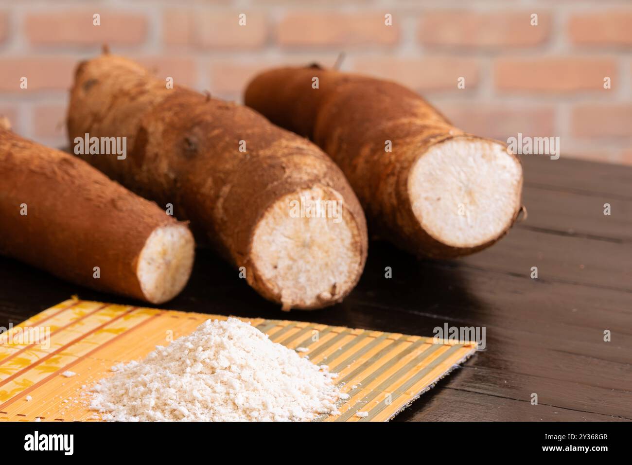 Una porzione di farina di manioca sopra un tappetino di bambù e radici di manioca sullo sfondo e fuori fuoco. Foto Stock