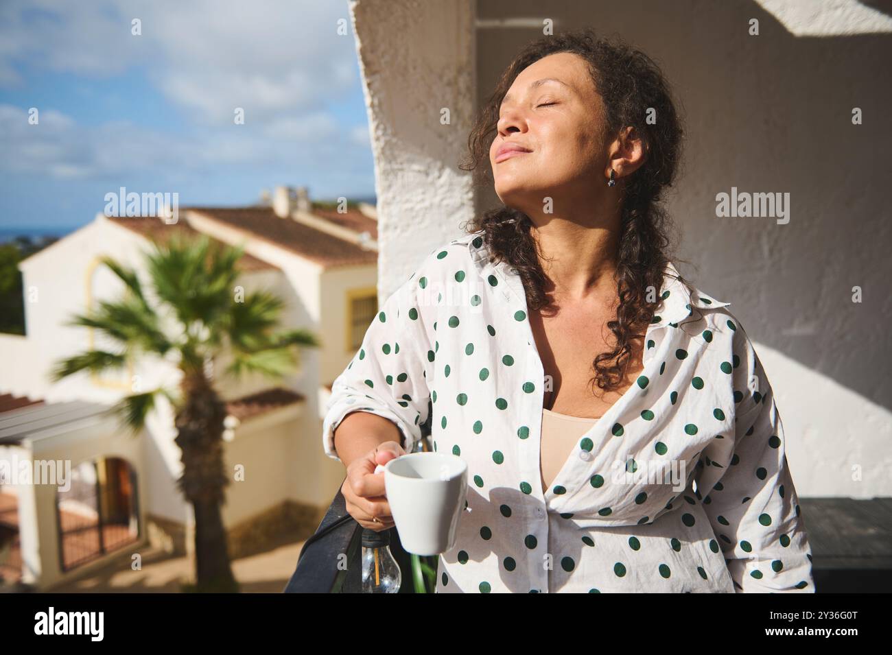 Una donna si crogiola al caldo sole del mattino su un balcone, assaporando il suo caffè e l'atmosfera tranquilla. Irradia soddisfazione e tranquillità. Foto Stock