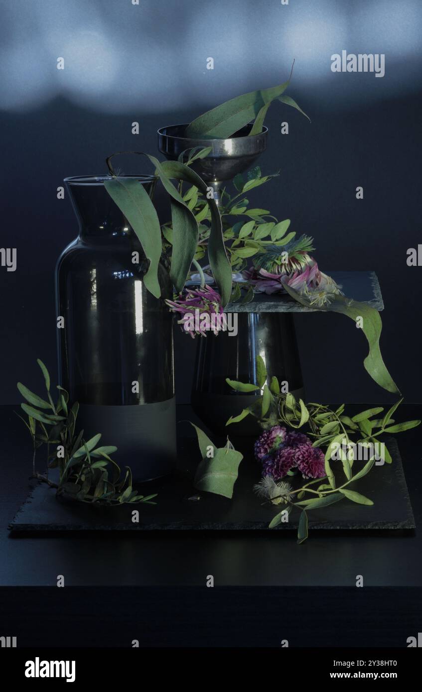 Fiori selvatici in vaso nero e vetro su sfondo scuro. Le foglie verdi essiccate aggiungono consistenza, creando un'atmosfera di decadimento. Foto Stock