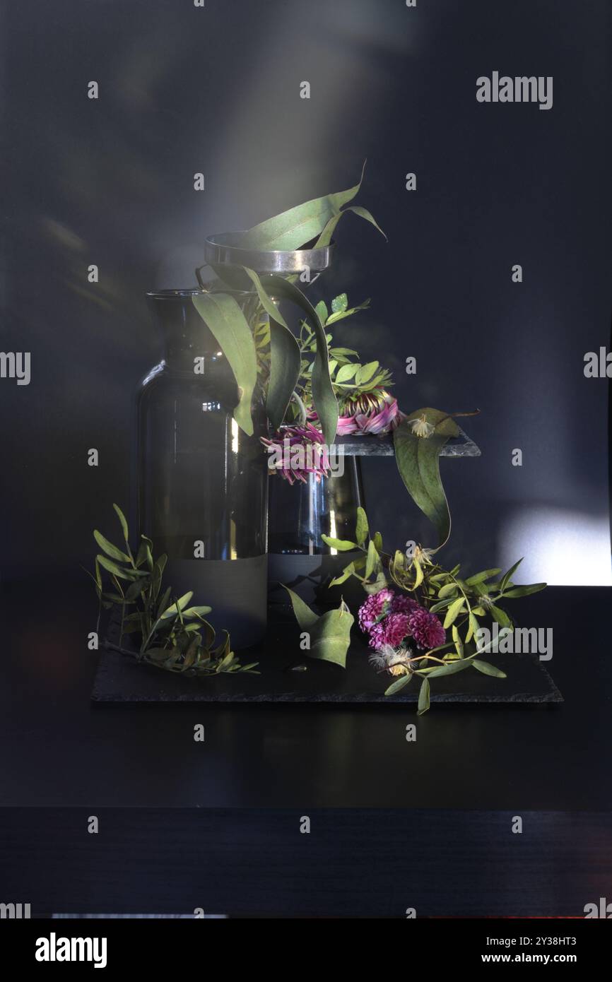 Vaso in vetro nero con fiori selvatici su sfondo scuro. Accanto ad esso, un bicchiere con foglie secche e fiori crea una natura morta profonda e drammatica. Foto Stock