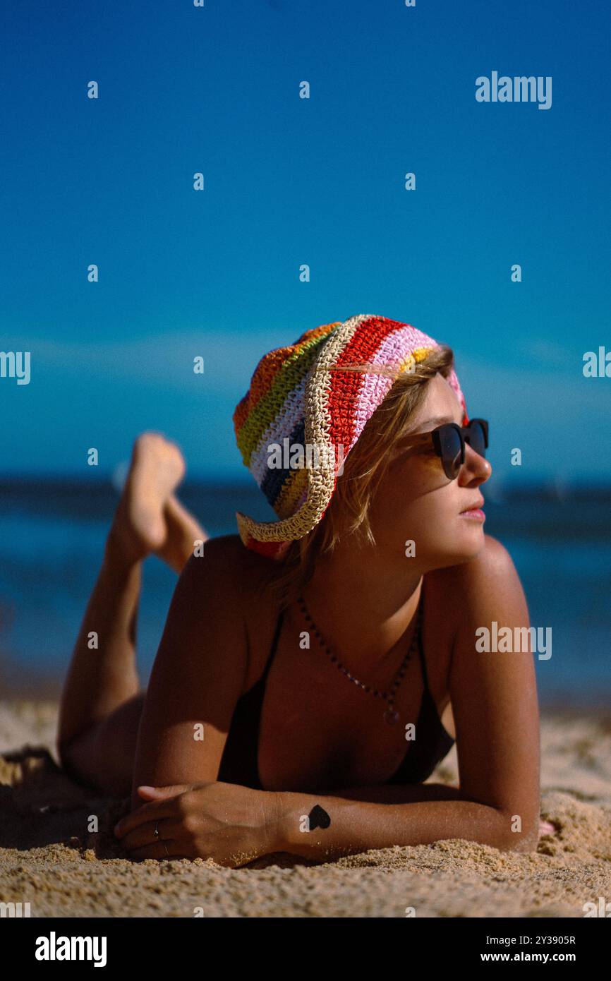 Una donna con costume da bagno e un cappello multicolore si appoggia sulla spiaggia. Foto Stock