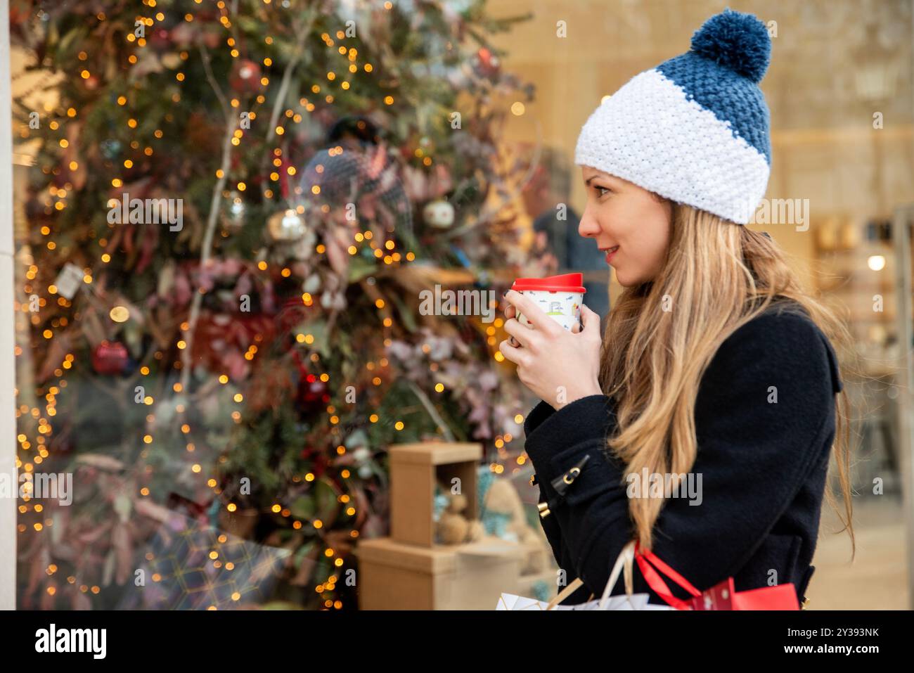 Donna con un cappello blu e bianco con una tazza di caffè rossa e borse di Natale, ammirando una vetrina festosa con un albero e luci Foto Stock