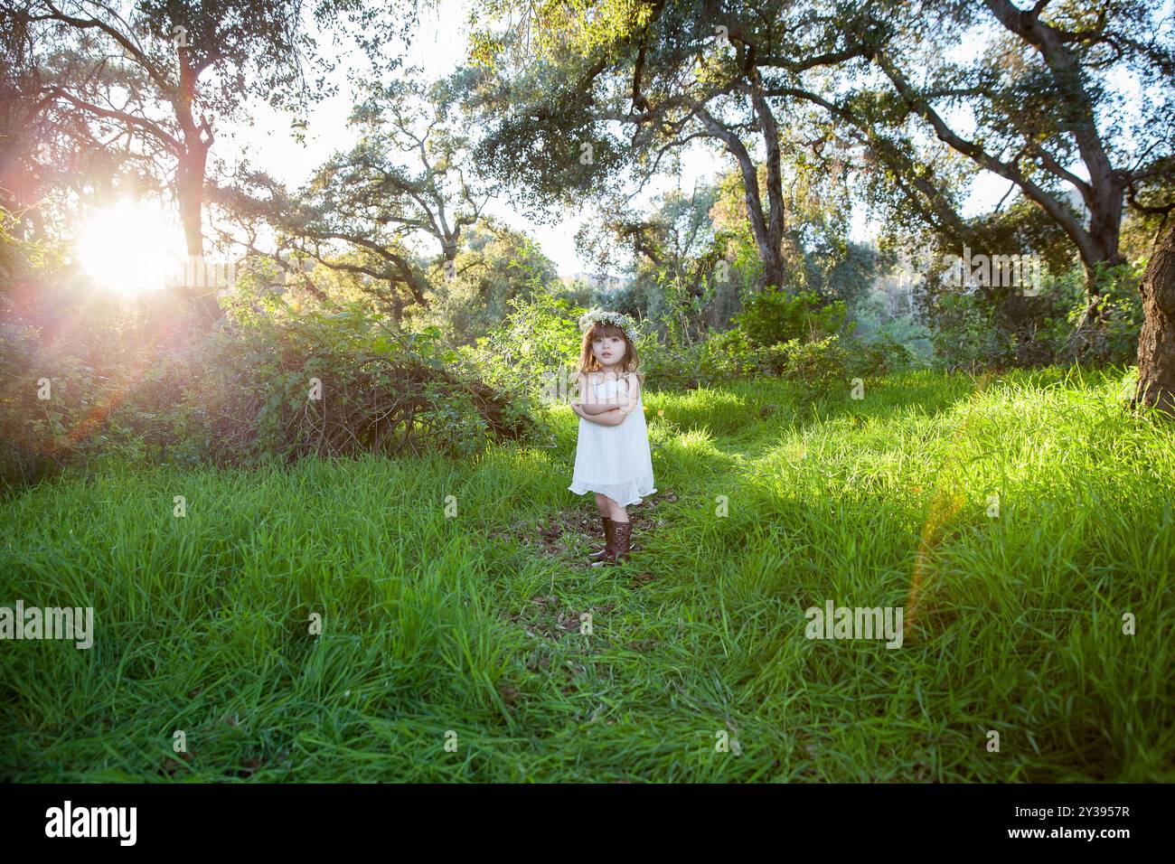 Bambino con un abito bianco e una corona floreale in piedi in una foresta illuminata dal sole Foto Stock