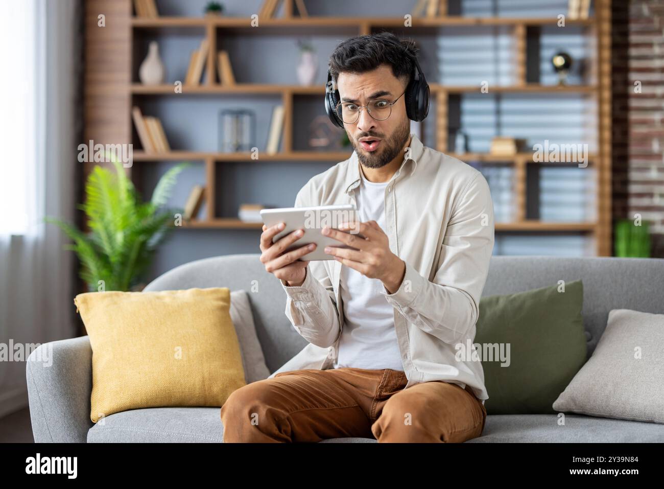 L'uomo con le cuffie è sorpreso mentre guarda film spaventosi sul tablet. Seduto sul divano, è assorbito dal contenuto, mostrando reazioni emotive. Foto Stock