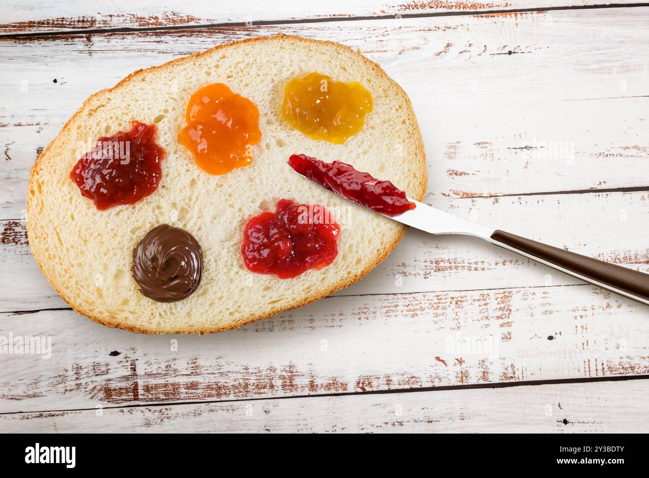 Fetta di pane bianco con vari tipi di marmellata e coltello. Vista dall'alto su un tavolo in legno. Foto Stock