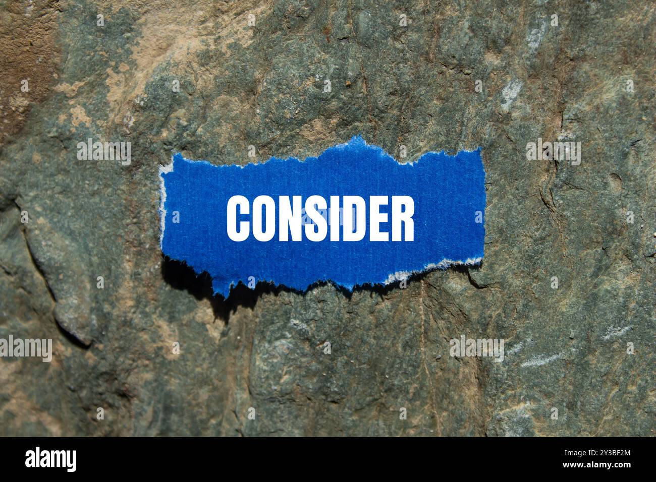 Considerare il messaggio scritto su un pezzo di carta blu strappato con sfondo in pietra. Simbolo concettuale da considerare. Copia spazio. Foto Stock