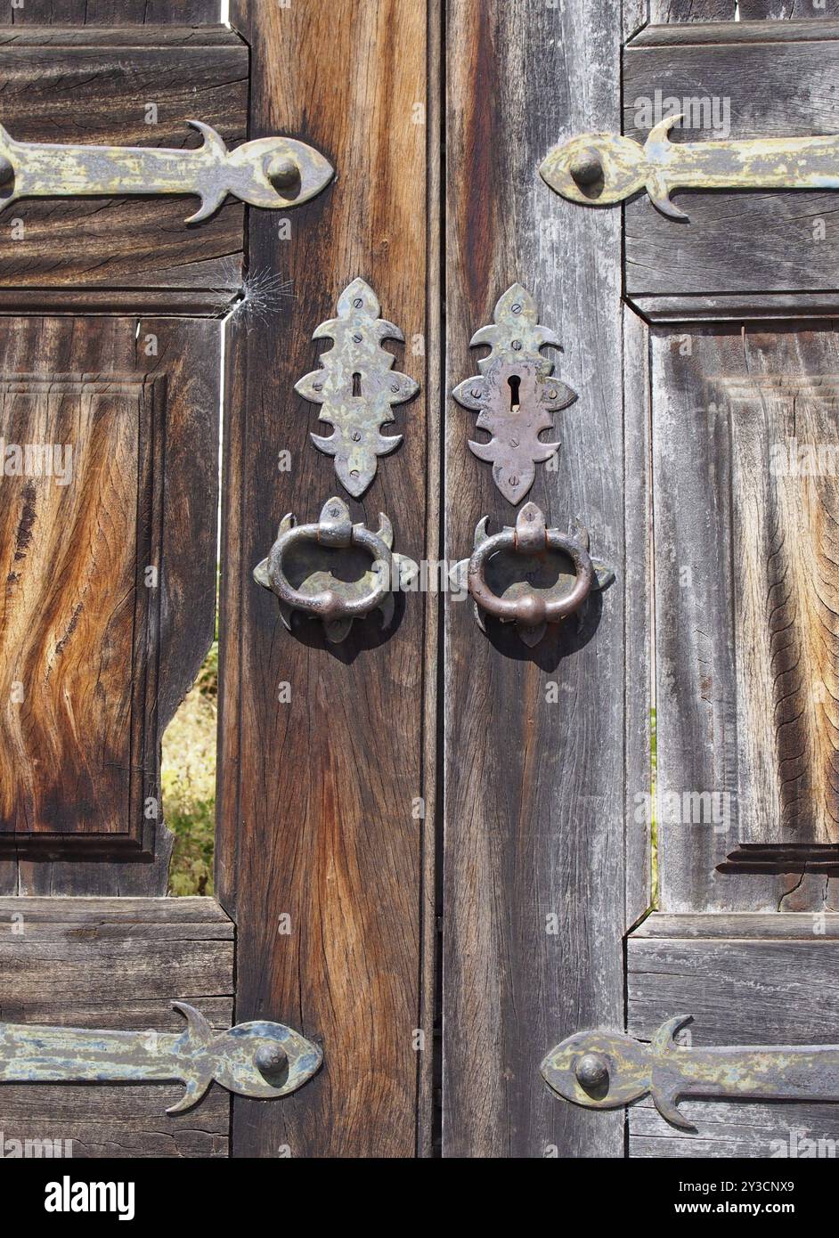 Un primo piano di vecchie doppie porte esterne in legno rotte e incrinate con maniglie ornate in ferro e ottone, fori e cerniere Foto Stock