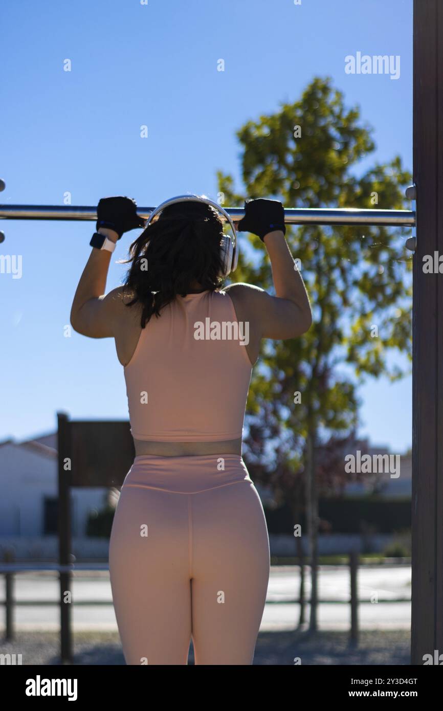 Ritratto verticale di una donna atletica che tira su barre parallele in una giornata di sole con cuffie e abbigliamento sportivo Foto Stock