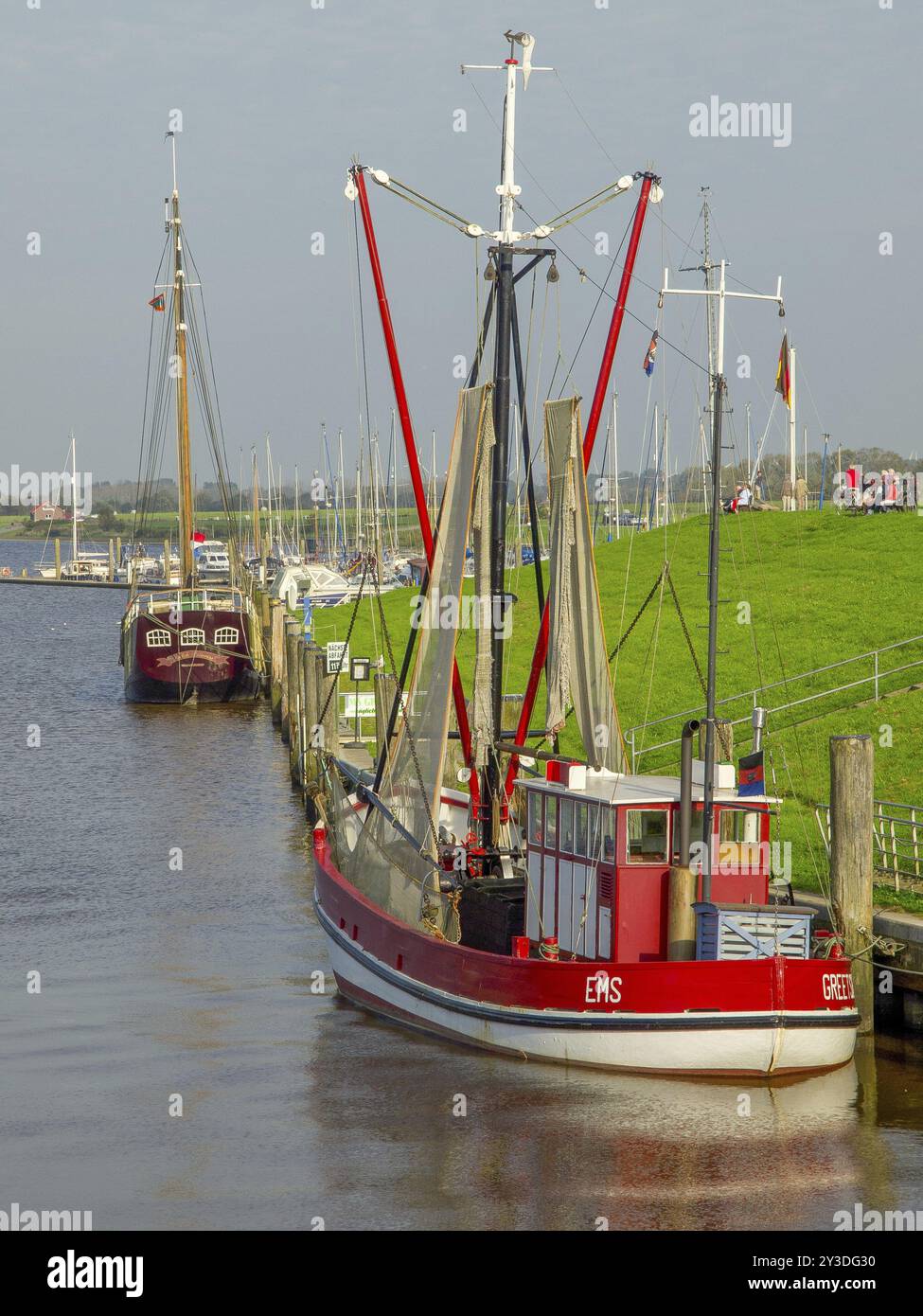 Un peschereccio rosso e bianco e un'altra barca sono ormeggiati in un molo nel porto vicino a un argine erboso, greetsiel, krummhoern, frisia orientale, ge Foto Stock