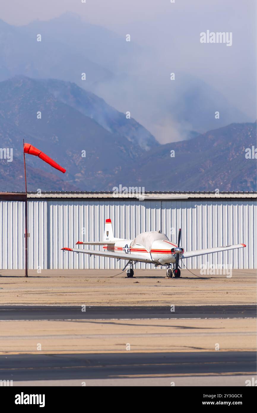 Un aereo privato ad ala bassa è legato vicino a un indicatore del vento, il che indica un'attività del vento moderata. Il fumo degli incendi boschivi è visibile su una montagna vicina sullo sfondo. Foto Stock
