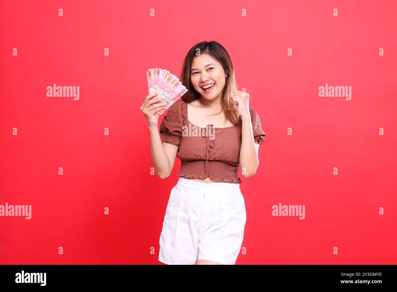 gesto di una allegra donna indonesiana che stringe il pugno con successo, tenendo in mano denaro la rupia indossando una camicetta marrone con sfondo rosso. per Foto Stock