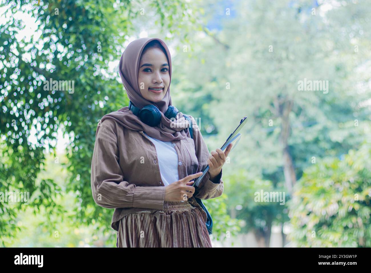 Giovane donna indonesiana in velo sorriso candid che porta libro e appunti in piedi in un parco all'aperto. studentesse per i temi dell'educazione, technolo Foto Stock