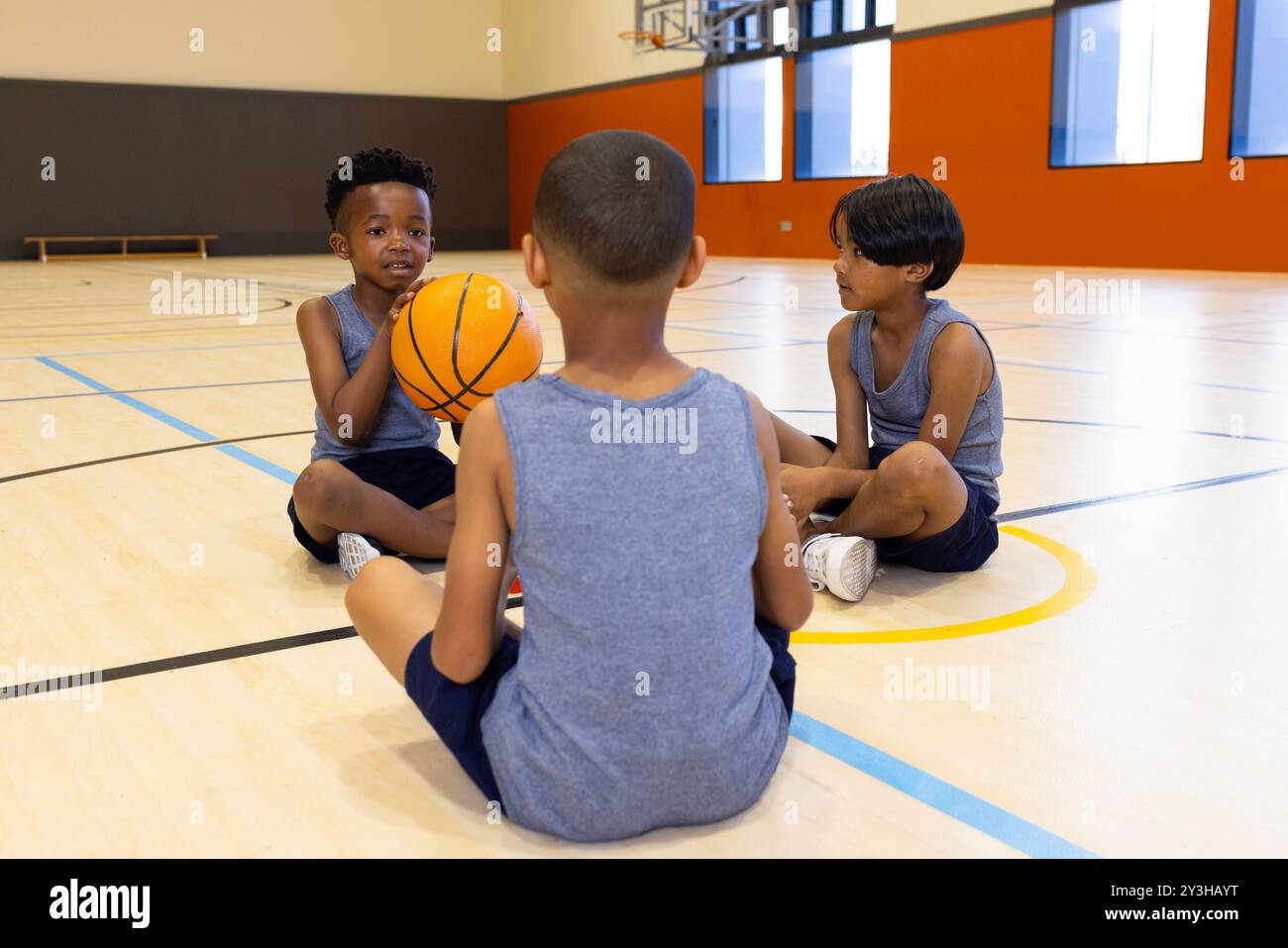Nella palestra scolastica, ragazzi multirazziali seduti sul pavimento che tengono il basket, parlano insieme Foto Stock