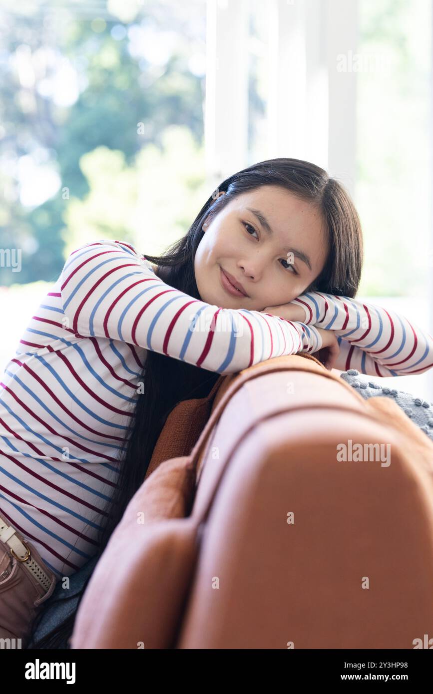 A casa, rilassatevi sul divano, giovane donna asiatica in camicia a righe sorridente e sognante ad occhi aperti Foto Stock