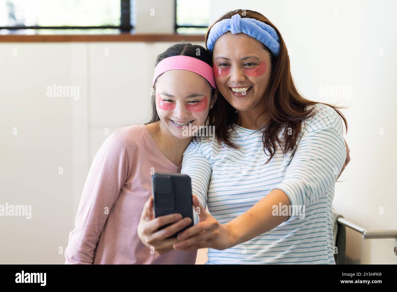 A casa, a fare selfie, madre asiatica e figlia sorridono con maschere facciali in bagno Foto Stock