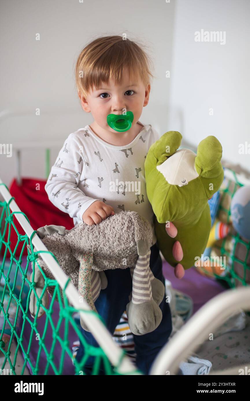 Un bambino allegro sorride mentre stringe un peluche verde e un morbido animale grigio, circondato da un'atmosfera giocosa in una vivace area giochi. Foto Stock